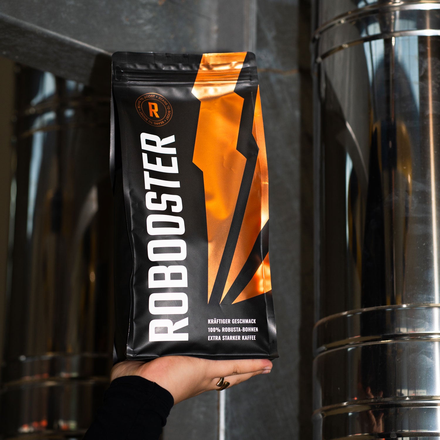 Eine Hand hält eine Robooster Kaffeepackungen mit ganzen Bohnen im dunklen Set-up.